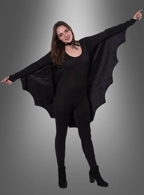 Eine Frau, die das „Fledermaus“-Cape für Damen und Herren trägt, steht mit ausgestreckten Armen da und präsentiert den flügelartigen Stoff vor einem grauen Hintergrund mit Farbverlauf – eine auffällige Wahl für ein Halloween-Kostüm für Damen.