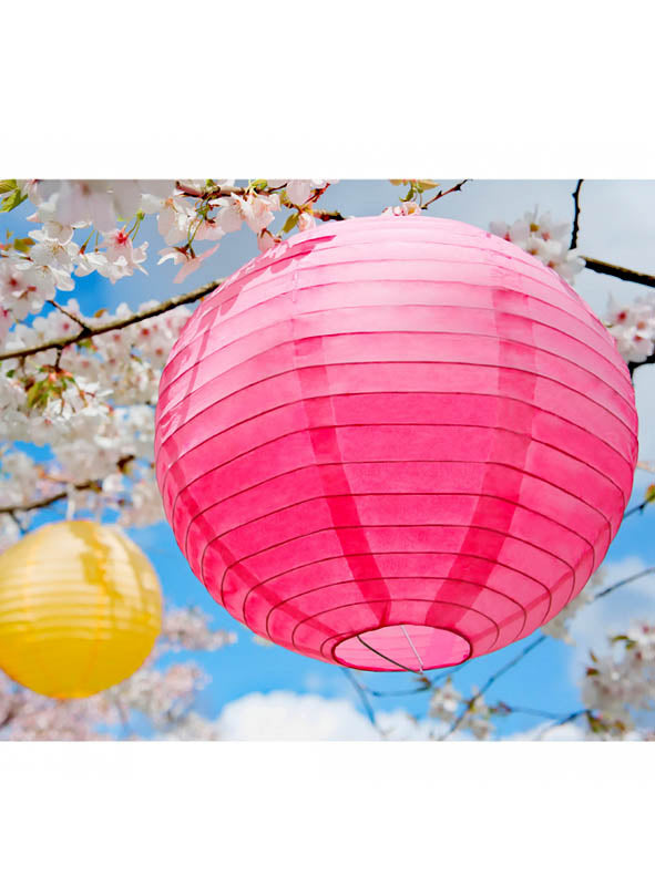 Eine leuchtend rosa Laterne aus der Kollektion „Papierlampions kräftige Farben 20–45 cm“ hängt an einem mit weißen Blüten geschmückten Baum und verleiht der Szene eine festliche Note; im Hintergrund runden ein gelber Papierlampion und der blaue Himmel diese bezaubernde Gartenparty-Dekoration ab.