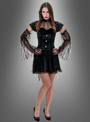 Eine Frau trägt das „Schwarze Vampir-Kleid Bella mit Spitze“, das sich durch elegante Spitzenärmel und einen Spitzenrock auszeichnet. Dazu trägt sie schwarze Pumps und dunkles Make-up und steht vor einem grauen Hintergrund mit Farbverlauf.
