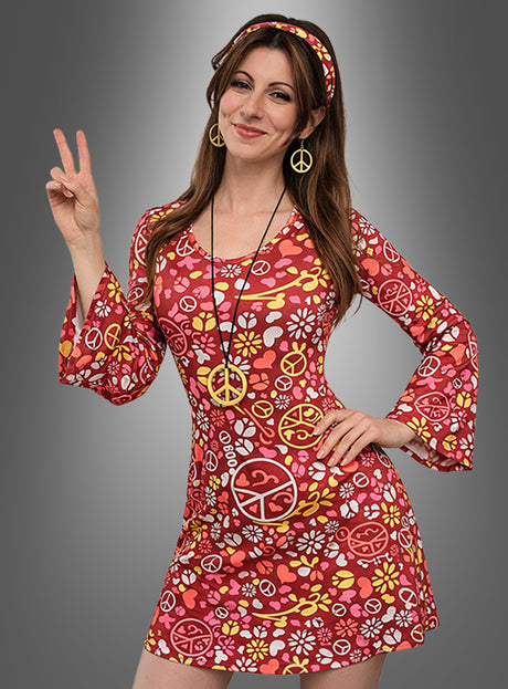 Eine Frau, gekleidet in das 5-teilige Hippie-Set mit Kleid – darunter ein rotes, mit Blumenmuster verziertes Kleid im Stil der 1960er Jahre, ein Stirnband und eine Halskette mit Peace-Zeichen –, posiert vor einem schlichten grauen Hintergrund, während sie mit der rechten Hand das Peace-Zeichen zeigt.