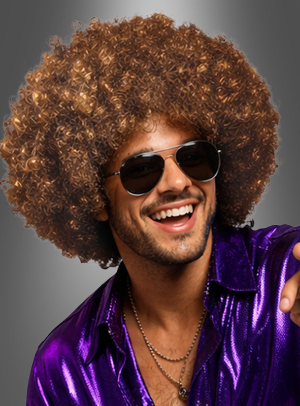 Ein Mann, der die „Super Riesen Afroperücke Deluxe Unisex“ trägt, dazu eine schwarze Sonnenbrille und ein glänzendes lila Hemd, lächelt vor einem grauen Hintergrund in die Kamera und verkörpert damit perfekt den klassischen 70er-Jahre-Kostüm-Look.