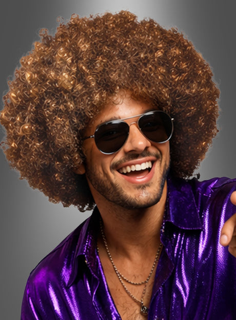Ein Mann, der die „Super Riesen Afroperücke Deluxe Unisex“ trägt, dazu eine schwarze Sonnenbrille und ein glänzendes lila Hemd, lächelt vor einem grauen Hintergrund in die Kamera und verkörpert damit perfekt den klassischen 70er-Jahre-Kostüm-Look.