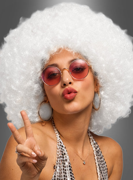 Eine Person, die eine „Super Riesen Afroperücke Deluxe Unisex“ in Weiß trägt, kombiniert mit einer runden, pinkfarbenen Sonnenbrille, großen Ohrringen und einem gemusterten Oberteil, macht vor einem grauen Hintergrund ein Peace-Zeichen und einen Kussmund – perfekt für jede 70er-Jahre-Kostümparty.