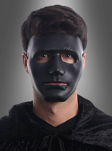 Eine Person trägt eine neutrale Gesichtsmaske in Schwarz oder Weiß und einen dunklen Umhang, steht vor einem grauen Hintergrund und blickt geradeaus.