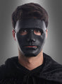 Eine Person trägt eine neutrale Gesichtsmaske in Schwarz oder Weiß und einen dunklen Umhang, steht vor einem grauen Hintergrund und blickt geradeaus.