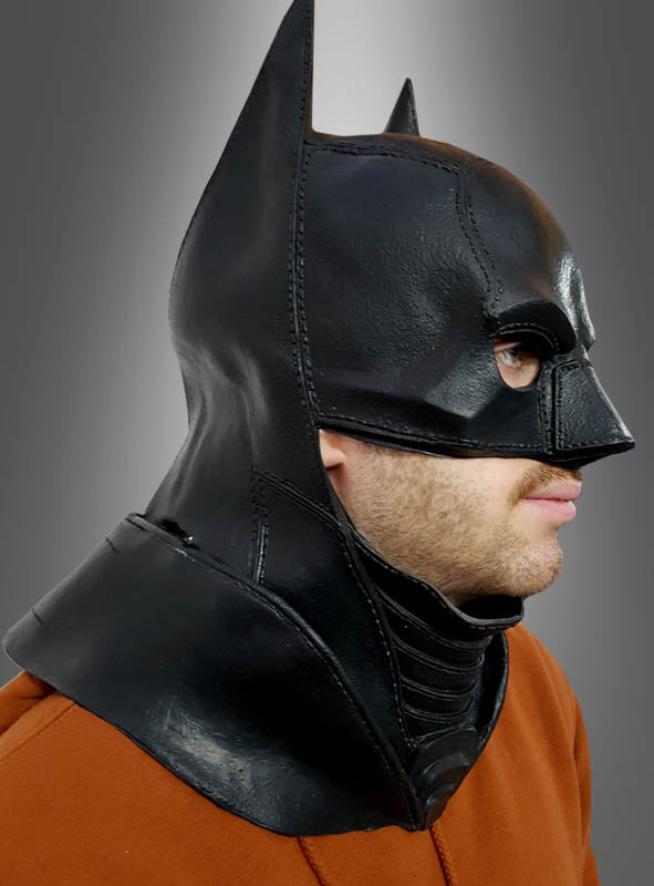 Eine Person trägt die Original DC Batman-Latexmaske für Erwachsene, die an die Maske aus „The Dark Knight Rises“ erinnert, sowie braune Kleidung; sie ist von der Seite vor einem grauen Hintergrund mit Farbverlauf zu sehen.