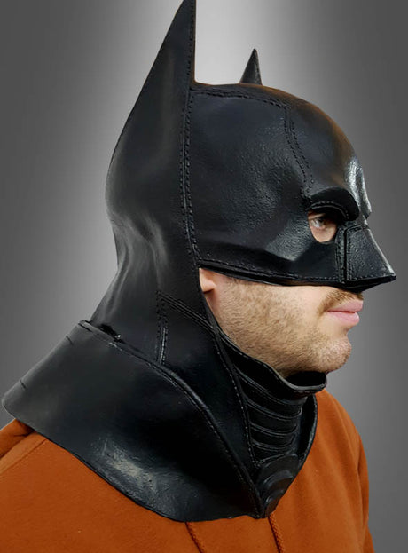 Eine Person trägt die Original DC Batman-Latexmaske für Erwachsene, die an die Maske aus „The Dark Knight Rises“ erinnert, sowie braune Kleidung; sie ist von der Seite vor einem grauen Hintergrund mit Farbverlauf zu sehen.