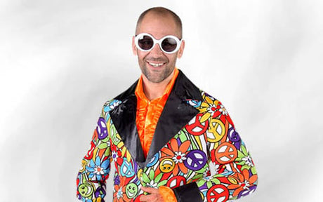 Ein Mann mit einer runden weißen Sonnenbrille und einem bunten Sakko mit Peace-Zeichen, Blumen und Smileys über einem orangefarbenen Hemd, der vor einem schlichten weißen Hintergrund steht.