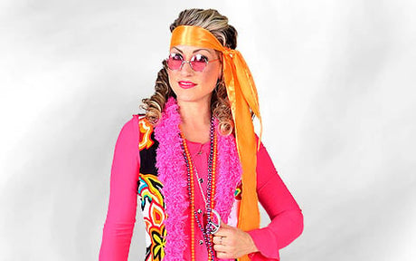 Eine Person in einem farbenfrohen, von den 1970er Jahren inspirierten Kostüm mit einem bunten Stirnband, einer rosa Sonnenbrille, einer gemusterten Weste und mehreren übereinander getragenen Halsketten vor einem einfarbigen Hintergrund.
