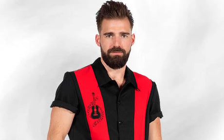 Ein bärtiger Mann, der ein schwarzes Hemd mit Kragen, roten Längsstreifen und einer Gitarrenstickerei trägt, steht vor einem schlichten, hellen Hintergrund.