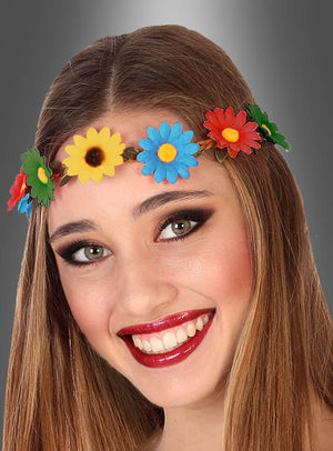 Eine junge Frau mit langen braunen Haaren lächelt vor einem grauen Hintergrund mit Farbverlauf und trägt dabei das von der Flower-Power-Party inspirierte Stirnband mit bunten Blumen.