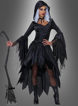 Eine Person, die ein schwarzes Glamour-Hexenkleid mit Kapuze trägt, dazu eine Kapuze, eine silberne Perücke und ausgestellte Ärmel, und die einen Besen in der Hand hält, steht vor einem grauen Hintergrund.