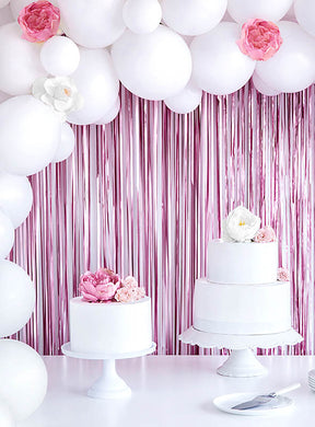 Auf Tortenständern stehen zwei weiße Torten mit rosa und weißen Blumen; im Hintergrund schimmert ein „Party Deko Lametta-Vorhang 250 cm“, der die festliche Dekoration perfekt ergänzt.
