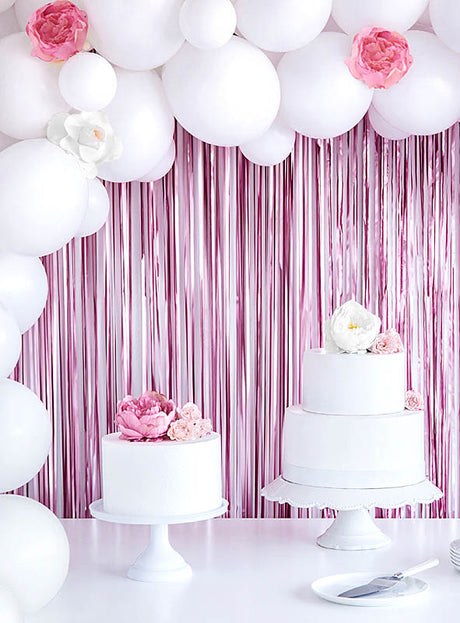 Auf Tortenständern stehen zwei weiße Torten mit rosa und weißen Blumen; im Hintergrund schimmert ein „Party Deko Lametta-Vorhang 250 cm“, der die festliche Dekoration perfekt ergänzt.