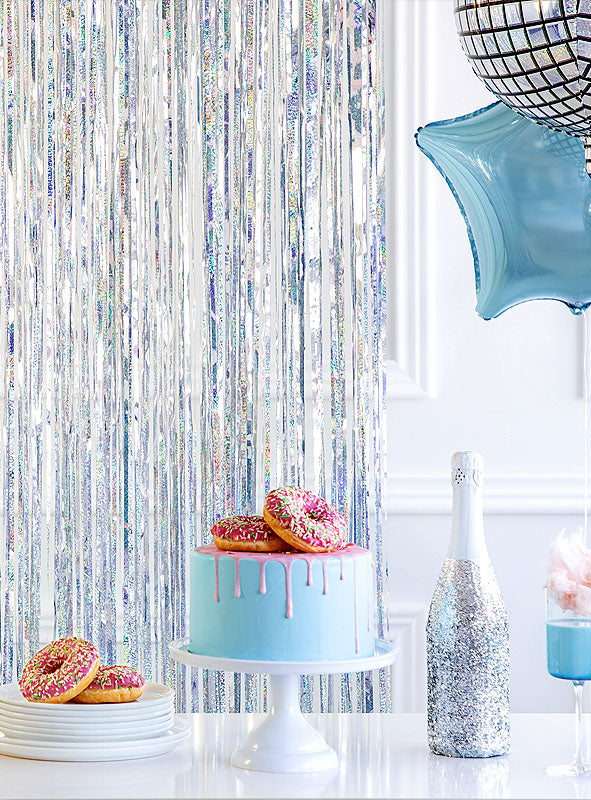 Auf einem Ständer steht eine blaue Torte mit rosa Zuckerguss und Donuts darauf, daneben eine glitzernde Flasche, Donuts, ein Sternballon, Zuckerwatte und ein silberner Lametta-Vorhang (250 cm) als Party-Dekoration.