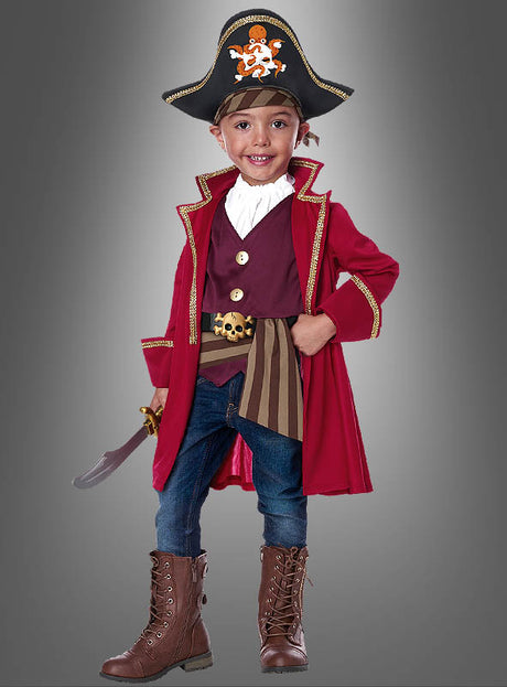Ein lächelndes Kind, das ein „Piratenkostüm Shorty für Kinder“ trägt – bestehend aus einem roten Mantel, braunen Stiefeln, einer gestreiften Schärpe und einem klassischen Piratenhut –, steht vor einem grauen Hintergrund und hält ein Spielzeugschwert in der Hand.
