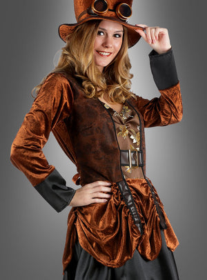 Eine Frau, die das Steampunk-Kleid „Kleid braun“ trägt, dazu einen passenden Hut und eine Schutzbrille, lächelt und posiert mit einer Hand in der Hüfte vor einem grauen Hintergrund.
