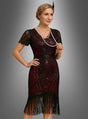 Eine Frau, die das „Flapperkleid Agatha im Stil der 20er Jahre“ trägt – ein schwarz-rotes, mit Fransen verziertes Kleid im Stil der 1920er Jahre mit Perlenstickerei und kurzen Ärmeln, ergänzt durch mehrreihige Perlenketten –, steht vor einem grauen Hintergrund mit Farbverlauf.