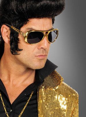 Ein Mann mit schwarzer Perücke, einer goldenen „King of Rock Elvis Brille 60er“-Sonnenbrille und einer goldenen Paillettenjacke über einem schwarzen Hemd mit Kragen blickt vor einem grauen Hintergrund zur Seite.