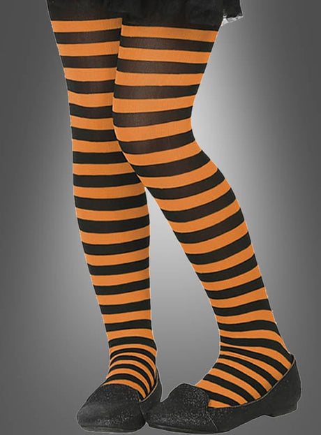 Eine Person, die orange-schwarze „Kinder Ringelstrumpfhosen“ und schwarze Schuhe trägt und vor einem grauen Hintergrund mit Farbverlauf steht.
