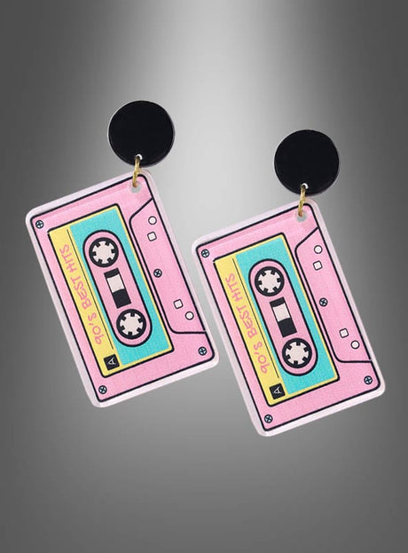 Zwei Ohrringe in Form von retro-rosa Kassetten mit der Aufschrift „90s BEST HITS“, befestigt an schwarzen, runden Steckelementen befestigt und vor einem grauen, verlaufenden Hintergrund präsentiert – diese verspielten Musik-Kassetten-Ohrringe im Stil der 80er- und 90er-Jahre sind ein perfektes Beispiel für angesagten Modeschmuck und erinnern an klassische Kassettendesigns der 80er Jahre.
