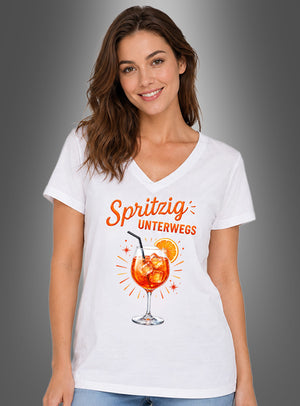 Eine Frau steht vor einem grauen Hintergrund mit Farbverlauf und trägt das Cocktail-T-Shirt „Spritzig unterwegs“ – ein weißes Shirt mit einem Aufdruck eines Aperol Spritz und dem Schriftzug „Spritzig unterwegs“ auf der Vorderseite. Dieses auffällige Gruppenshirt „Sommer“ eignet sich perfekt für Sommerausflüge und Treffen bei warmem Wetter.