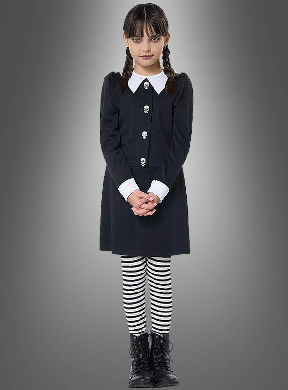 Ein Mädchen im „Dunkles Gothic Mädchen Kostüm“ – bestehend aus einem schwarzen Kleid mit weißem Kragen und Manschetten, gestreiften Leggings und schwarzen Stiefeln – steht vor einem grauen Hintergrund mit Farbverlauf, blickt mit gefalteten Händen nach vorne und ist damit ideal für alle, die auf der Suche nach einem authentischen „Wednesday Kostüm Kinder“-Look sind.