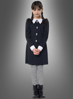 Ein Mädchen im „Dunkles Gothic Mädchen Kostüm“ – bestehend aus einem schwarzen Kleid mit weißem Kragen und Manschetten, gestreiften Leggings und schwarzen Stiefeln – steht vor einem grauen Hintergrund mit Farbverlauf, blickt mit gefalteten Händen nach vorne und ist damit ideal für alle, die auf der Suche nach einem authentischen „Wednesday Kostüm Kinder“-Look sind.