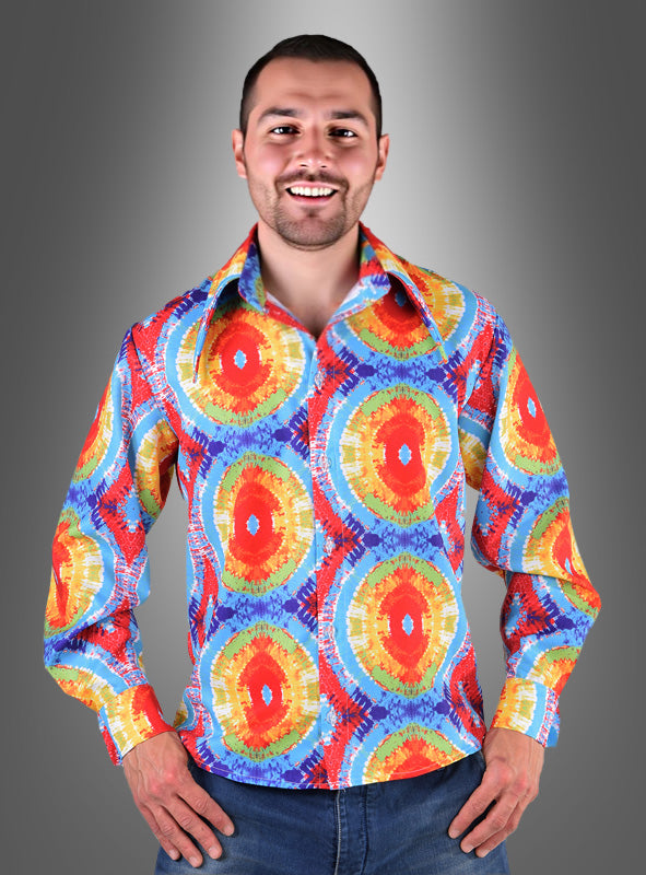 Ein lächelnder Mann, der ein buntes Hippie-Hemd im Batik-Stil für Herren und blaue Jeans trägt, steht vor einem grauen Hintergrund mit Farbverlauf.