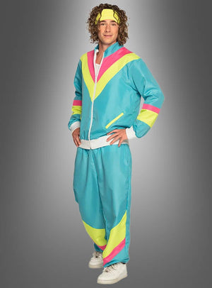 Eine Person, die den „80er Trainingsanzug blau“ trägt – einen Retro-Trainingsanzug im Stil der 80er Jahre in Blau, Gelb und Rosa – mit einem passenden Stirnband und weißen Sneakers, vor einem grauen Hintergrund mit Farbverlauf.