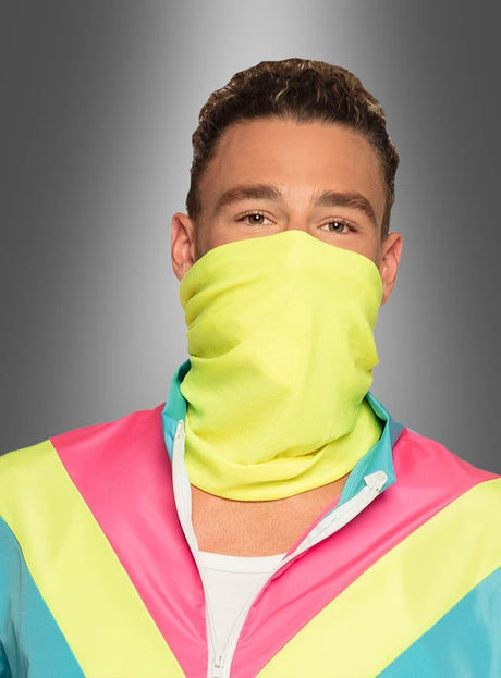 Ein Mann, der einen neongelben Mund-Nasen-Schutz und den „80er Trainingsanzug blau“ mit leuchtend pinken, türkisfarbenen und gelben Streifen trägt, posiert vor einem grauen Hintergrund mit Farbverlauf.
