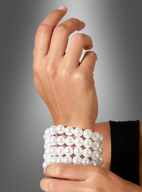 Eine Person, die das „Deluxe Perlenarmband 20er Jahre weiß“ trägt – ein breites Armband mit drei Reihen großer weißer Perlen –, bewundert das elegante Accessoire mit einer Hand und verkörpert damit den opulenten Stil der Kostüme der 20er Jahre und den Reiz von hochwertigem Kostümschmuck.