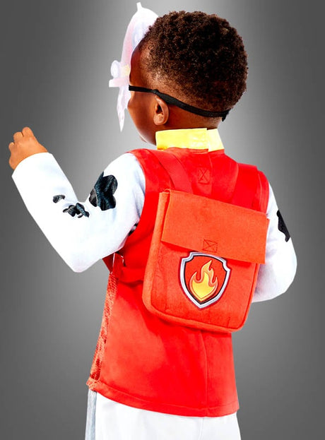 Ein Kind in einem roten Marshall-Kostüm „Paw Patrol Deluxe Hund“ mit einem Flammen-Emblem und einem Ärmel mit Pfotenabdruck steht mit dem Rücken zum Betrachter vor einem grauen Hintergrund.