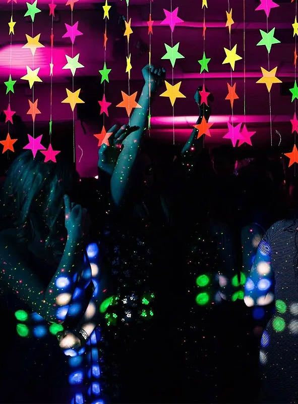 Die Partygäste tanzen unter Neon-Stern-Dekorationen und leuchtenden Lichtern, ihre Silhouetten und erhobenen Hände heben sich deutlich vom dunklen Hintergrund ab – all dies wird durch die auffälligen 80er-Jahre-Neon-Girlanden (440 cm) und die UV-leuchtende Girlande unterstrichen und sorgt so für die ultimative Schwarzlicht-Party-Dekoration.