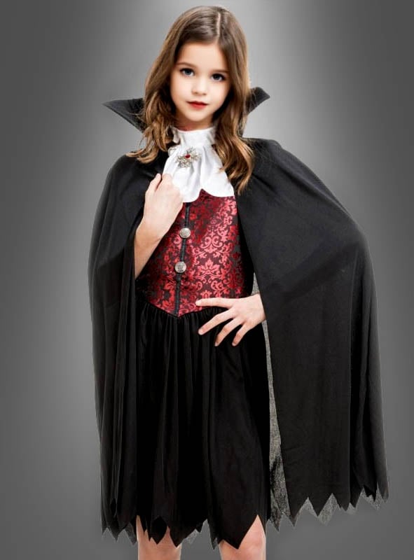 Ein junges Mädchen steht selbstbewusst vor einem grauen Hintergrund und trägt das „Vampirin-Kostüm Theodora für Mädchen“ – ein schwarzes Outfit mit einer roten Weste, einem weißen Rüschenkragen und einem hochgeschlossenen Umhang.
