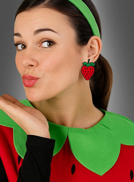 Eine Frau, die ein Kostüm mit Erdbeer-Motiv und Erdbeer-Ohrringe (Hänger oder Stecker) aus der Sommer-Modeschmuck-Kollektion trägt, dazu einen grünen Kragen und ein Stirnband, wirft der Kamera einen Kuss zu.