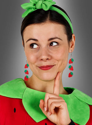 Eine Frau mit einem grünen Stirnband, Erbeeren-Ohrringen (Hänger oder Stecker) als Obstschmuck und einem Kostüm mit grünem Kragen und rotem Stoff, die mit dem Finger am Kinn zur Seite blickt.