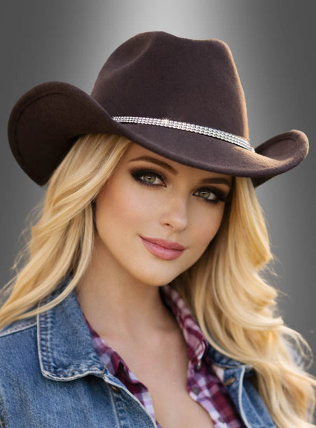 Eine Frau mit langen blonden Haaren trägt den „Cowboy Hut mit Strass Deluxe“ – einen braunen Westernhut, der mit einem Strass-Hutband verziert ist – kombiniert mit einer Jeansweste und einem karierten Hemd, und posiert vor einem grauen Hintergrund mit Farbverlauf.