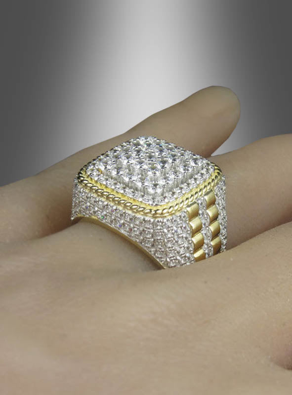Ein „Goldring mit Strass für Herren“ – mit einem quadratischen, mit Diamanten besetzten Oberteil in eleganten Gold- und Silbertönen – wird an einem Finger vor einem neutralen Hintergrund getragen.