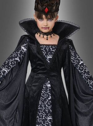 Ein Kind, das das „Schwarze Vampir-Königin“-Kinderkostüm „Lina“ trägt – ein dramatisches schwarz-weißes Vampir-Königin-Kostüm für Kinder mit auffälligem Kragen, Krone und einem schwarzen Vampir-Kleid –, steht vor einem grauen Hintergrund; für einen zusätzlichen Effekt wurde ihm Kunstblut um den Mund herum aufgetragen.