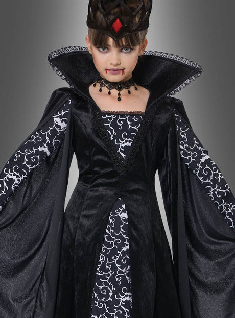 Ein Kind, das das „Schwarze Vampir-Königin“-Kinderkostüm „Lina“ trägt – ein dramatisches schwarz-weißes Vampir-Königin-Kostüm für Kinder mit auffälligem Kragen, Krone und einem schwarzen Vampir-Kleid –, steht vor einem grauen Hintergrund; für einen zusätzlichen Effekt wurde ihm Kunstblut um den Mund herum aufgetragen.