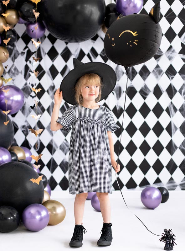 Ein junges Mädchen in einem karierten Kleid und einer Mütze mit Katzenohren steht inmitten der „Glossy Balloons Chrome Partydeko“, umgeben von metallisch schimmernden schwarzen, violetten und goldenen Dekorationen vor einer festlichen Halloween-Kulisse.