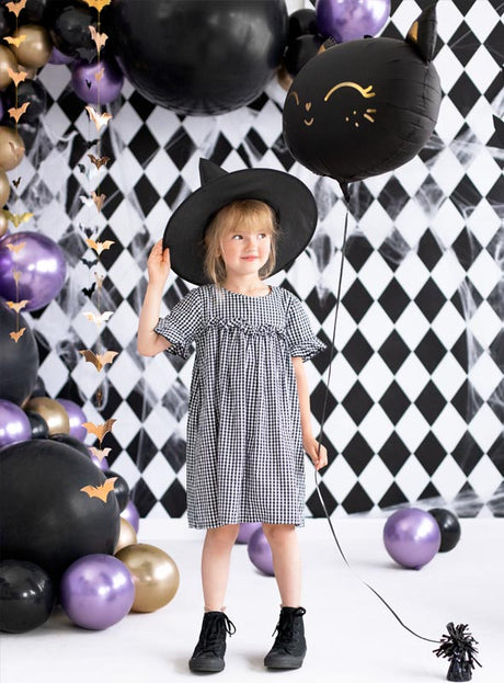 Ein junges Mädchen in einem karierten Kleid und einer Mütze mit Katzenohren steht inmitten der „Glossy Balloons Chrome Partydeko“, umgeben von metallisch schimmernden schwarzen, violetten und goldenen Dekorationen vor einer festlichen Halloween-Kulisse.