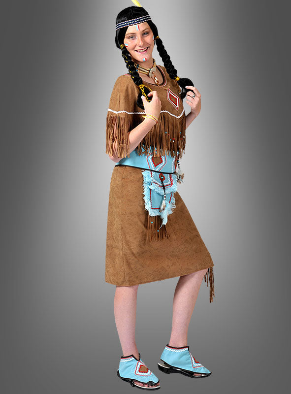 Eine Person, die das Kostüm „Die furchtlose Squaw“ trägt – bestehend aus einem braunen Kleid im Wildleder-Look mit Fransenverzierung, geflochtenen Haaren, einem mit Perlen bestickten Stirnband, einem blauen Gürtel und passenden blauen Schuhen –, posiert vor einem grauen Hintergrund mit Farbverlauf.