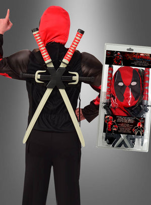 Eine Person, die ein rot-schwarzes Deadpool-Kostüm mit Kapuze und zwei Spielzeugschwertern auf dem Rücken trägt, steht neben einem verpackten Deadpool-Kostüm-Set mit Waffen, das eine Maske und Waffenzubehör enthält.
