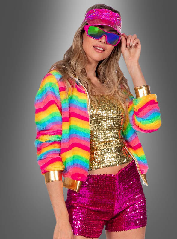 Eine Frau posiert selbstbewusst vor einem grauen Hintergrund und trägt dabei die „Kurze Plüschjacke Regenbogen“ mit leuchtenden Regenbogenstrifen, kombiniert mit einem goldenen Pailletten-Top, rosa Pailletten-Shorts, einer rosa Schirmmütze und einer reflektierenden Sonnenbrille.