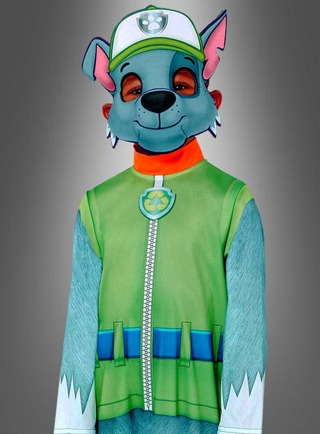 Ein Kind trägt das Rocky-Kostüm aus „Paw Patrol“ in den Farben Grün und Blau mit passender Mütze und Maske und steht vor einem grauen Hintergrund mit Farbverlauf.