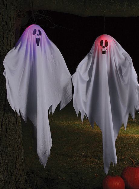 Zwei „Lustige Geist-Dekos mit Licht, 50 cm“ – eine leuchtet violett, die andere rot – hängen nachts unter einem Baum und tauchen die Szene in ein gespenstisches Licht. Diese beleuchteten Geisterfiguren sorgen für eine eindrucksvolle Halloween-Dekoration, während orangefarbene Kürbisse in der Nähe die gruselige Atmosphäre vervollständigen.