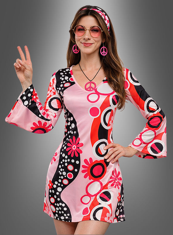 Eine Frau, die das rosa „Hippie-Kleid“ mit den Accessoires „Dream Circles“ trägt und dazu ein passendes Stirnband sowie Schmuck mit Peace-Zeichen kombiniert hat, posiert mit ausgestrecktem Peace-Zeichen vor einem grauen Hintergrund mit Farbverlauf – damit ist dies das ideale Mottoparty-Outfit für jede Veranstaltung im Retro-Stil.