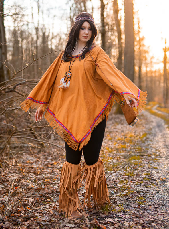 Eine Frau steht auf einem Waldweg und trägt einen hellbraunen Poncho mit Fransen, Hippie-Stulpen mit Fransen und ein gemustertes Stirnband, während das Sonnenlicht durch die Bäume fällt.